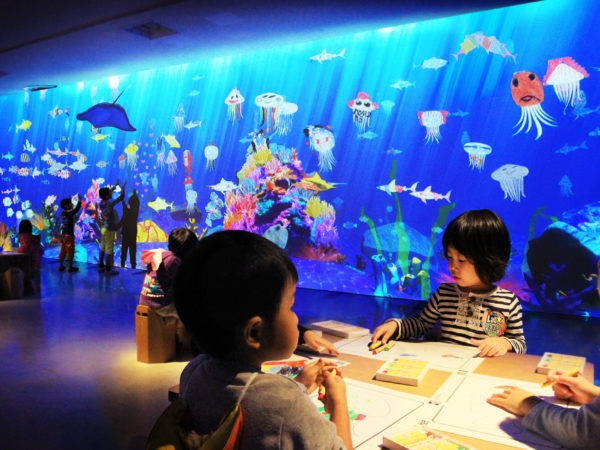 Children color pictures at a table in a room with walls covered in projections of animated sea creatures.